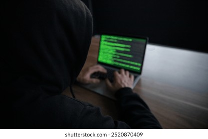 A person in a dark hoodie types on a laptop displaying green code. The image represents themes of hacking, cybersecurity, digital security, and online anonymity in a low-light environment.