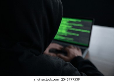 A person in a dark hoodie types on a laptop displaying green code. The image represents themes of hacking, cybersecurity, digital security, and online anonymity in a low-light environment.