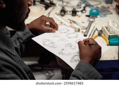 Persona diseñando intrincadas piezas de joyería en una mesa artesanal llena de varias herramientas y materiales creando obras de arte, mientras está sentado en un taller rodeado de creatividad y artesanía