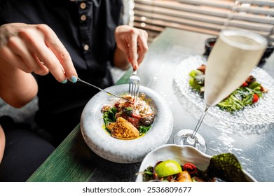 A person dining, focusing on their hands using utensils to eat a gourmet dish, with a glass of champagne nearby. Elegant table setting, fine dining experience, close-up shot - Powered by Shutterstock