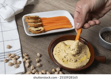 A person is dipping a carrot into a bowl of hummus. The bowl of hummus is on a table next to a plate of bread