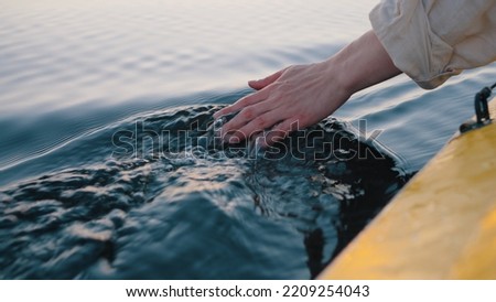 Person dips hand in calm river water sitting in yellow boat. Man enjoys camping and sailing on boat. Guy makes waves on water with hand
