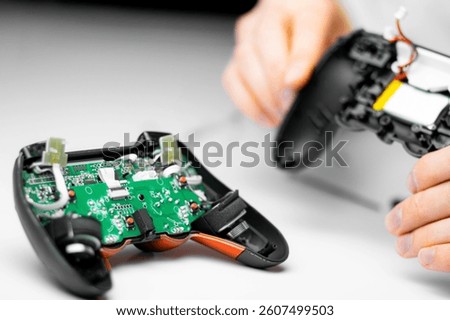 A person disassembling a black gaming controller, exposing internal green circuit boards and wiring. The hands hold the separated shell on a white background.

