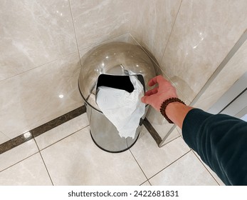 A person is discarding a roll of toilet paper into a trash can on the bathroom floor, made of composite material and hardwood flooring.