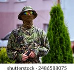 A person dressed in camouflage clothing and a face covering stands in front of a lush evergreen tree, holding a firearm. They are wearing a wide-brimmed hat and sunglasses, with a serious expression 