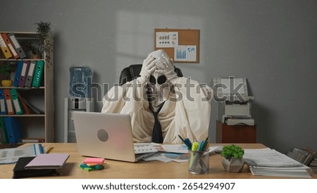 Person dressed as ghost day at work. Halloween businessman ghost in white sheet necktie sitting at office desk works with financial papers, tired expression holding head. Halloween business concept.