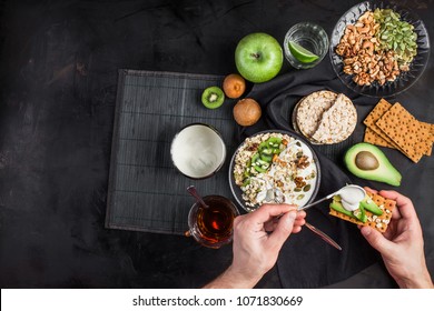 A Person Eats Eating Flakes With Yogurt. Useful Food In The Hands. A Healthy Breakfast All Day Long. A Bowl With Cereals, Nuts And Yogurt. Glass Of Water On A Dark Background. Useful Food 2