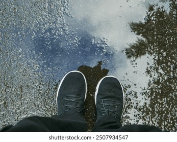 A person feet in dark jeans and shoes standing on wet asphalt road, cloudy blue sky and green trees reflection in puddle. Travel and tourism concept. Outdoor scene. - Powered by Shutterstock