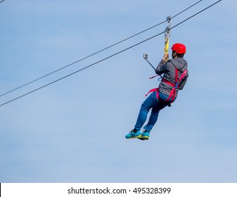 Person Flying Under A Zip Line. He Is Taking A Selfie On The Way Across. His Back Is Facing The Camera So His Face Is Not Visible. The Sky Above Is White And Blue. 