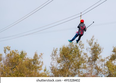 Person Flying Under A Zip Line. He Is Taking A Selfie On The Way Across.  His Back Is Facing The Camera So His Face Is Not Visible. The Sky Above Is Hazy, Trees Are In The Background.