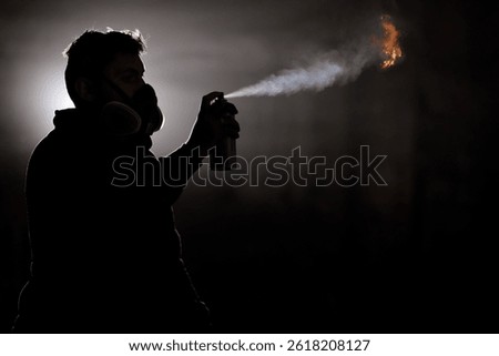 A person in a gas mask sprays paint into the air, creating a dramatic effect with a fiery burst. The dark surroundings enhance the intensity of the scene during nighttime creativity.