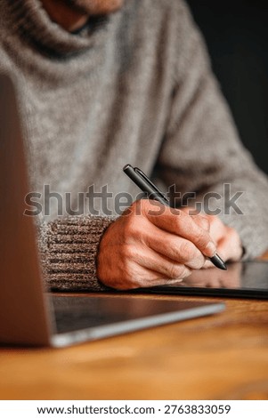 A person in a gray sweater is using a tablet and pen while working on a laptop, focusing on digital design.