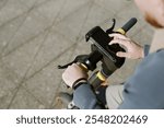 Person gripping handlebars of electric scooter while interacting with smartphone. Urban pavement visible in background, showing modern transportation method