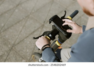 Person gripping handlebars of electric scooter while interacting with smartphone. Urban pavement visible in background, showing modern transportation method - Powered by Shutterstock