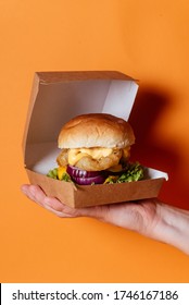 Person Hand Holding Chicken Meat Grilled Burger With White Golden Buns In Paper Box On Orange Background
