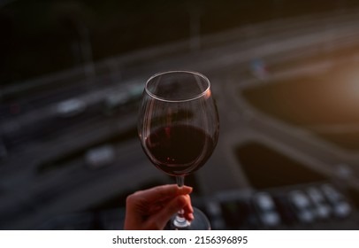  Person Hand Holds A Red Wine Alcohol Drink In A Wineglass Against City. No People. Copy Space