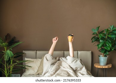The person has woken up at home from his bed and is lying under a warm blanket with a cup of coffee in the air. Difficulties with getting up early. Everyday affairs. Humorous