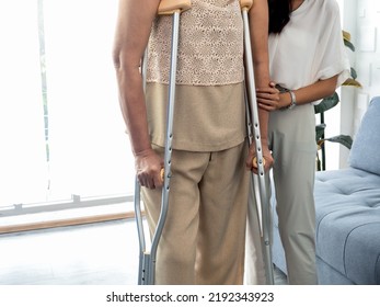 Person Helping, Female's Hand Hold The Arms Of An Elderly Woman Trying To Walk On Crutches Standing In Recovery Room Background. Helping And Care Senior Patient To Walk With Crutches Walker Concept.