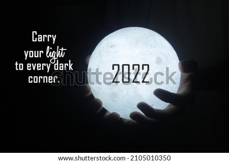 Person holding 2022 light of moon light in hands on dark black background. With positive inspirational motivational message - Carry your light to every dark corner. Be the light of hope concept.