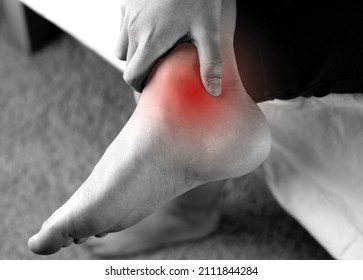  A person holding Achilles tendon by hands, suffering with pain in red spot area. Sprain ligament or Achilles tendonitis symptoms. Image in black and white with red highlights, Health care concept