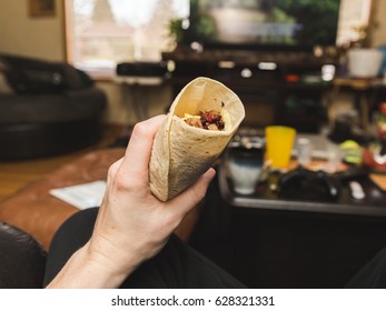 Person Holding Up A Breakfast Burrito While In A Living Room.