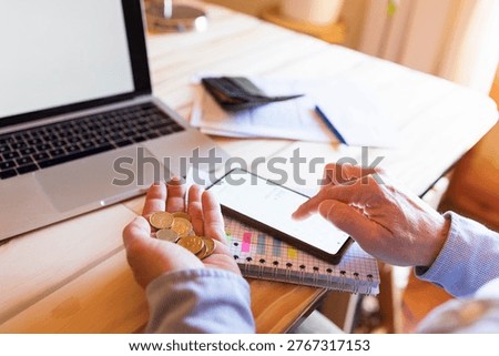 Person holding coins while checking finances on a smartphone, illustrating budgeting, counting money, rising costs, debt pressure and personal finance stress at a home desk