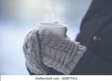 A person is holding a cup of hot drink outdoor. The weather is very cold and the mug is smoking. The person has a jacket and wool mittens in her hands. Image has vintage effect applied.