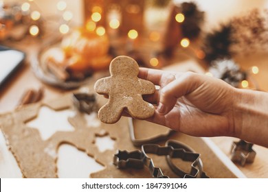 Person holding gingerbread man cookie, Christmas holiday advent. Hand cutting gingerbread dough with christmas man metal cutter on background of festive lights, decorations on rustic table