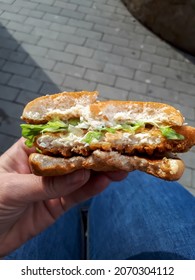 Person Holding In A Hand Half Eaten Burger With Chicken Patty, Shredded Lettuce And Creamy Mayonnaise In Toasted Bun Outdoors