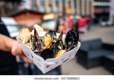 Person holding a paper container of cooked mussels with bread slices, in an urban outdoor setting. Ideal for street food and seafood cuisine themes - Powered by Shutterstock