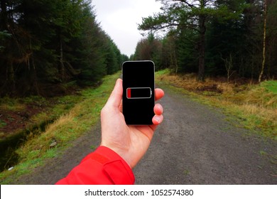 A Person Holding A Phone In Their Hand With Dead Battery. It Is Cold And Wet And In The Background A Trail Runs Through Coniferous Forest.