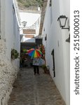 Person holding a rainbow umbrella walking down a narrow cobblestone alley with white walls and potted plants, Alpujarras, granada, andalucia, espa a