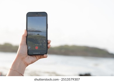 A person holding a smart phone in front of a scenic ocean view, capturing the landscape. The phone screen displays the same view, showing waves and a clear sky. - Powered by Shutterstock