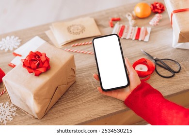 A person holding a smartphone amidst holiday gift wrapping materials, featuring a red ribbon bow on a gift package. The scene conveys a festive and creative atmosphere. - Powered by Shutterstock