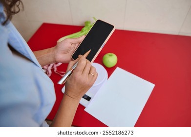 A person holding a smartphone and pen, recording health or diet-related data on a red counter beside a green apple. Organized planning for a balanced lifestyle is represented. - Powered by Shutterstock