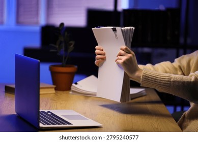 Person holding stack of documents with laptop on desk, business administration and office work concept, a person holds a large stack of white documents with a binder clip