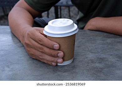 A person holding a takeaway coffee cup with a white lid, sitting at a table. This simple moment reflects relaxation, coffee culture, and a break from routine, perfect for leisure or outdoor café vibes - Powered by Shutterstock