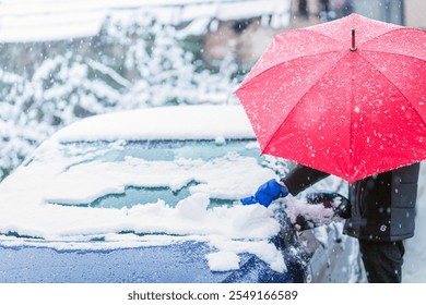 A person holding a vibrant red umbrella clears snow from a car windshield during a snowfall. The scene highlights a snowy day and the process of winter car maintenance. - Powered by Shutterstock