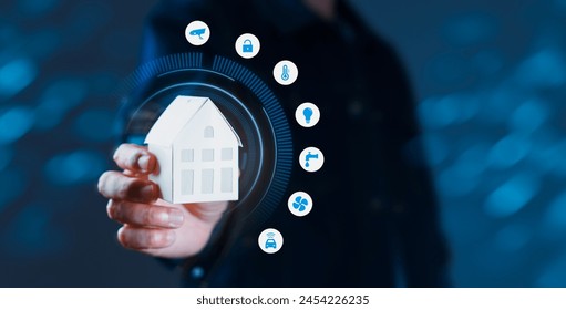 A person is holding a white house in their hand, with a blue background. The house is surrounded by various icons, including a car, a light, a clock, and a camera. Concept of a smart home