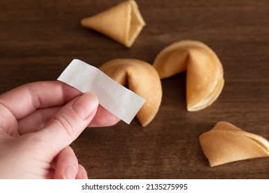 Person Holds In Hand Blank Paper Slip From Fortune Cookie Against Few Cookies Laying On Table Surface Background, Mockup For Your Good Luck Wish