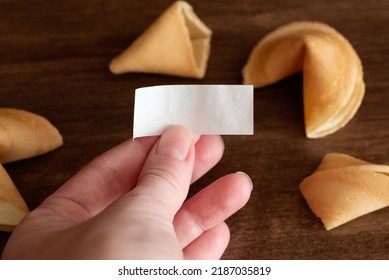 Person Holds In Hand Blank Paper Slip From Fortune Cookie Against Few Cookies Laying On Table Surface Background, Mockup For Your Good Luck Wish