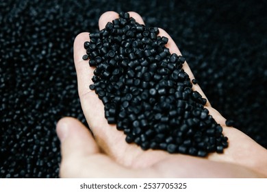 A person holds a handful of black plastic granules, showcasing the material's texture and color amidst a backdrop of similar pellets in a factory environment. - Powered by Shutterstock