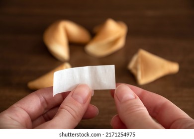 Person Holds In Hands Blank Paper Slip From Fortune Cookie Against Few Cookies Laying On Table Surface Background, Mockup For Your Good Luck Wish