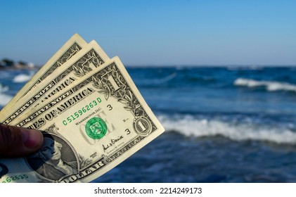 Person Holds In His Hand Three Dollar Bills On The Background Of The Waves Of The Blue Sea. Concept Money Finance Business Tourism Travel, Money Spending, Cash Expenses. Money Cash Expenses Business