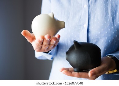 Person holds two piggy banks, white is smaller and lighter, black is bigger and heavier, like savings vs debt