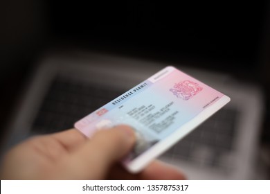 Person holds UK Residence Permit - BRP card in hand and computer in the background. Immigration concept image. 