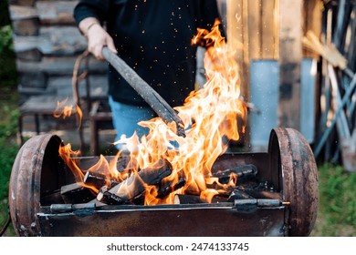 A person ignites a barbecue grill by placing wood logs and stoking the flames to achieve optimal heat for grilling. The scene captures the intensity and anticipation of outdoor BBQ cooking. - Powered by Shutterstock