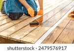 A person kneeling on a wooden deck applies a coat of wood stain with a roller. The wood is mostly bare, but some of the deck has already been stained, giving it a darker hue.