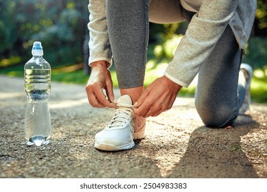 A person is kneeling outside in nature, diligently tying their running shoe, with a water bottle conveniently placed nearby, perfectly illustrating a healthy and active lifestyle they embrace daily - Powered by Shutterstock