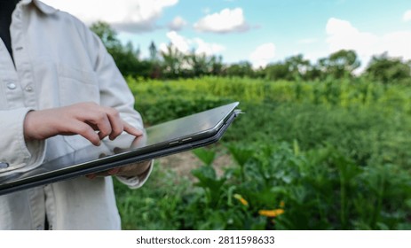 A person in a light-colored jacket holds a tablet and touches the screen while standing in a lush green field with crops and trees in the background under a cloudy sky